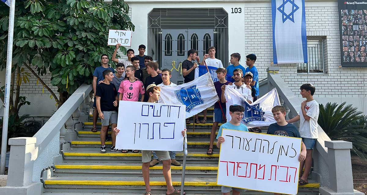 קבוצת נערים עומדים על מדרגות מחוץ לבניין עיריית רעננה עם שלטים ודגלי תנועת בני עקיבא. שני נערים בחזית מחזיקים שלטים עם הכיתובים: "דורשים פתרון" ו"למען תלמידי אורבך הבית מתחורר". ברקע ניתן לראות את מבנה הסניף של בני עקיבא עם דגל ישראל תלוי על הקיר.