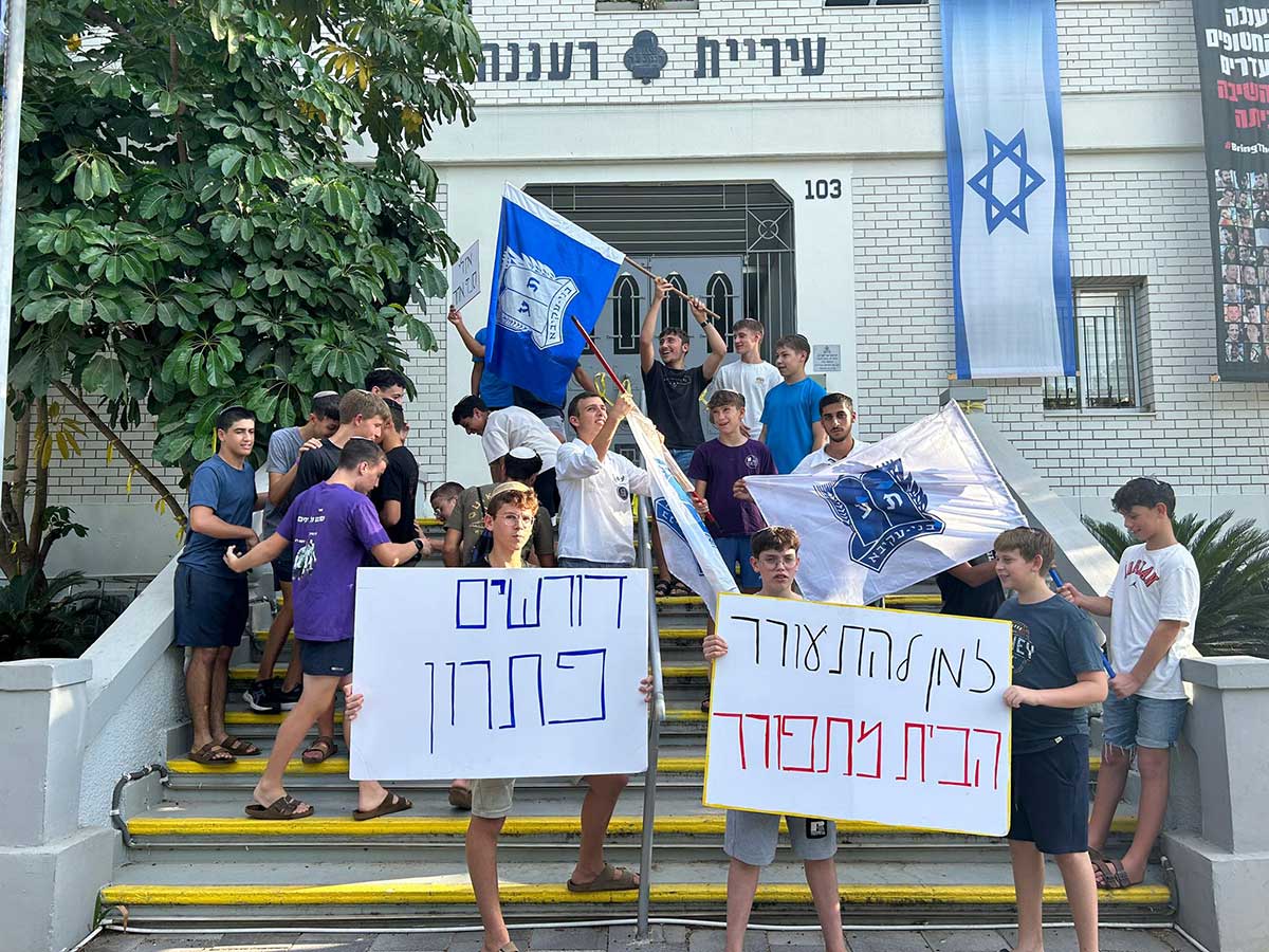קבוצת נערים עומדים על מדרגות מחוץ לבניין עיריית רעננה עם שלטים ודגלי תנועת בני עקיבא. שני נערים בחזית מחזיקים שלטים עם הכיתובים: "דורשים פתרון" ו"למען תלמידי אורבך הבית מתחורר". ברקע ניתן לראות את מבנה הסניף של בני עקיבא עם דגל ישראל תלוי על הקיר.
