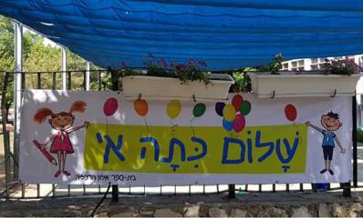 שלט צבעוני עם איורים של ילדים, המברך את תלמידי כיתה א' בבית הספר אלון בהרצליה. הכיתוב על השלט: "שלום כיתה א'". השלט מעוטר בבלונים צבעוניים ואיורים של ילדה וילד מחייכים.