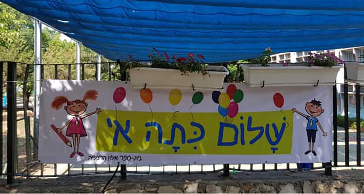 שלט צבעוני עם איורים של ילדים, המברך את תלמידי כיתה א' בבית הספר אלון בהרצליה. הכיתוב על השלט: "שלום כיתה א'". השלט מעוטר בבלונים צבעוניים ואיורים של ילדה וילד מחייכים.