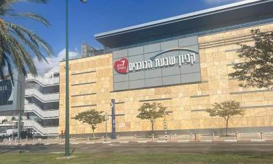 קניון שבעת הכוכבים, צילום חוץ של הצד המערבי של הקניון, הבניין מצופה באבן בצבע חול ובמרכז שלט כדול על המבנה: קניון שבעת הכוכבים, 7 ימים בשבילך. בצד שמאל במדרכה שלט פירסומת ועץ דקל