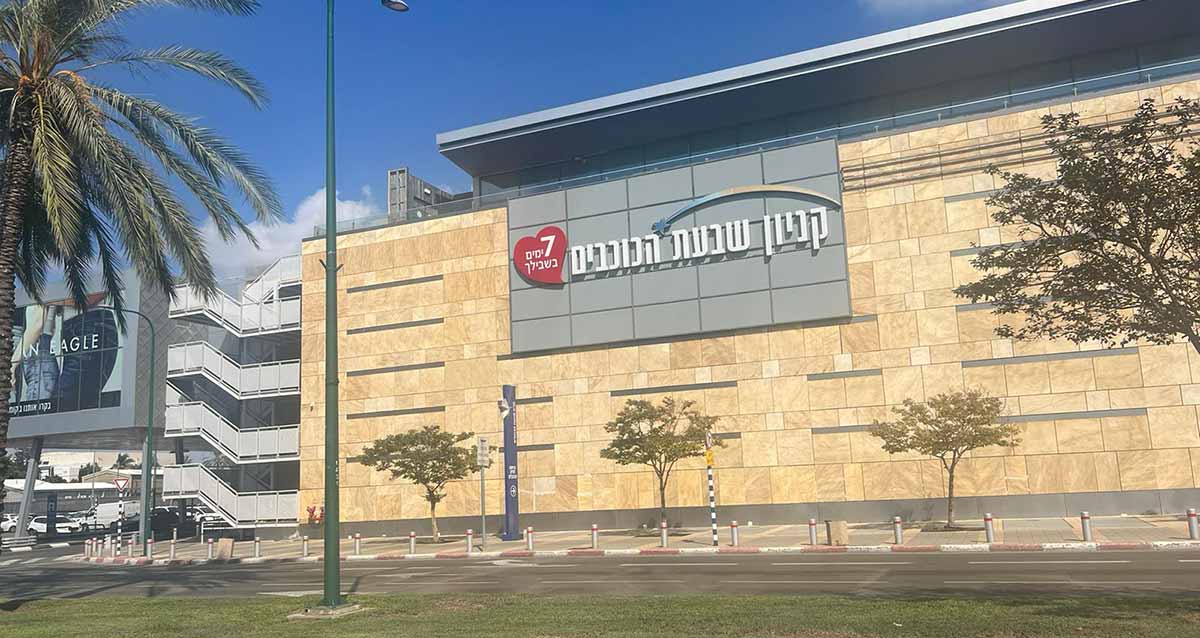 קניון שבעת הכוכבים, צילום חוץ של הצד המערבי של הקניון, הבניין מצופה באבן בצבע חול ובמרכז שלט כדול על המבנה: קניון שבעת הכוכבים, 7 ימים בשבילך. בצד שמאל במדרכה שלט פירסומת ועץ דקל