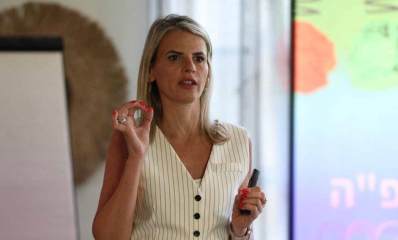 צילום פלג גוף עליון של שלומית שפיגלבלט, שיער בלונדיני לבושה בחולצת נטולת שרוולים מפוספסת מדברת ומחזיקה מצביע במהלך מצגת או הרצאה. ברקע ניתן לראות מסך עם כיתוב מטושטש, ככל הנראה כחלק מהמצגת שהיא מעבירה.