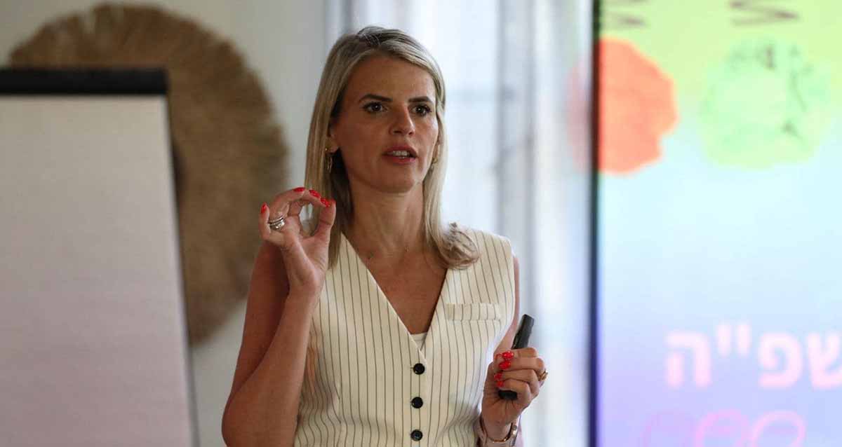 צילום פלג גוף עליון של שלומית שפיגלבלט, שיער בלונדיני לבושה בחולצת נטולת שרוולים מפוספסת מדברת ומחזיקה מצביע במהלך מצגת או הרצאה. ברקע ניתן לראות מסך עם כיתוב מטושטש, ככל הנראה כחלק מהמצגת שהיא מעבירה.