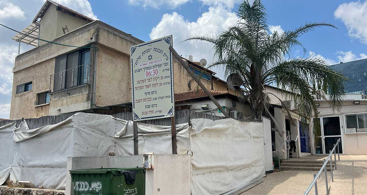 מבנה עם שלט על חזיתו המציין את שעות התפילות בבית הכנסת "מנוחת שלום". השלט מכיל את זמני התפילות ביום חול ובשבת, ומספר טלפון ליצירת קשר. השלט מעוטר במסגרת דקורטיבית ובכיתוב שחור על רקע לבן. בצד ימין של המבנה יש עץ גבוה ובחזית הכניסה רמפה ומדרגות המובילות פנימה. על השלט כתוב: בס"ד בית הכנסת "מנוחת שלום" זמני התפילות במניין השבועי שחרית: 06:30 מנחה 20 דקות לפני השקיעה ערבית 20 דקות לאחר השקיעה ביום שישי מנחה קבלת שבת וערבית 10 דקות לאחר הדלקת נרות שבת ביום שבת שחרית: 07:30