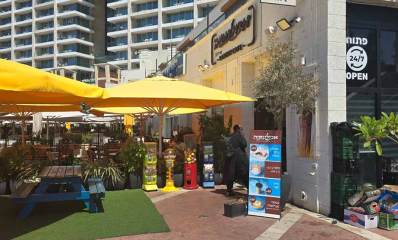 סעדת אבולעפיה בהרצליה, תמונה של השטח החיצוני, מימין מבנה של המסעדה ומשמאל בחוץ רחבת ישיבה עם שמשיות צהובות, שולחן וספסלי גינה בצבע כחול על דשא סינתטי, בצד שמאל מאחורי שורת עציצים ארוכים עם שיחים ואחוריהם מספר רב של שולחנות וכסאות ואנשים יושבים, הכל בחוץ. בצד ימין על המדרכה שלט ארוך ובחלק העליון כתוב: אבולעפיה באותיות לבנות על רקע חום. מתחת לכיתוב יש ארבע תמונות של מנות המסעדה ובצידן כיתוב המתאר את המנות.