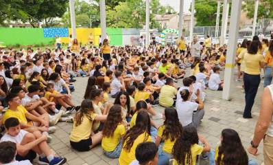 פתיחת שנת הלימודים בחטיבת אלון כפר סבא, צילום מהצד של התלמידים היושבים על הרצפה המרוצפת של חצר בית הספר. רבים מהתלמידים לובשים חולצות צהובות. בצד ימין של התמונה מורות ומורים גם חלק מהם בחולצות צהובות. ברקע גדרות חצר בית הספר, וצד אחד של הגדר מכוסה בלוחות צבעוניים.