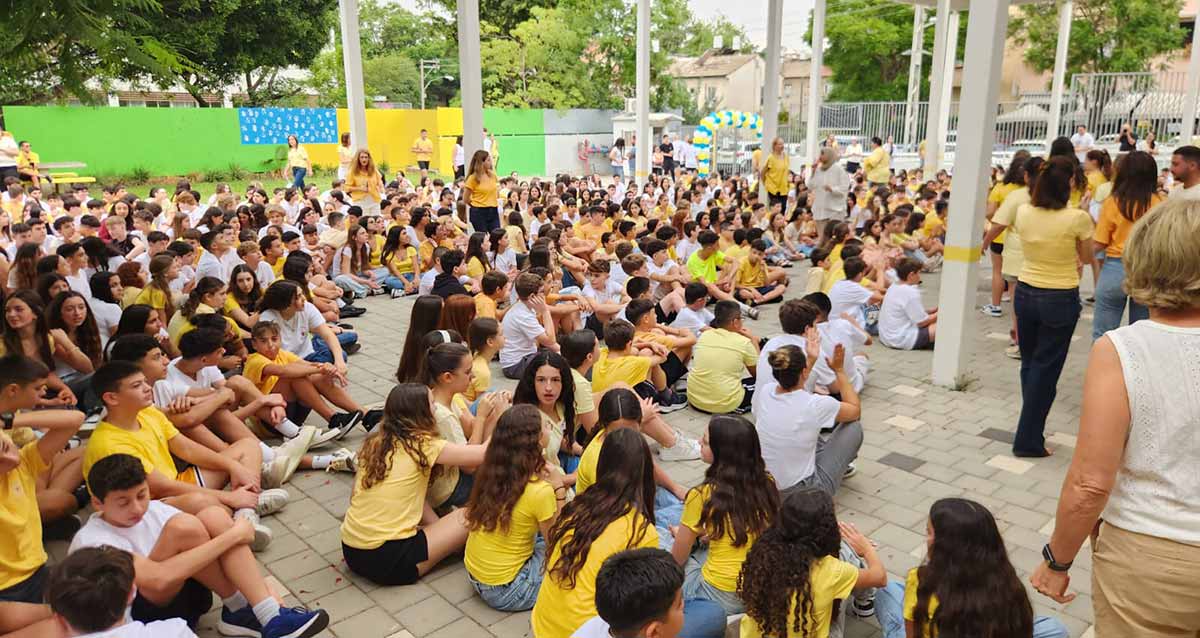 פתיחת שנת הלימודים בחטיבת אלון כפר סבא, צילום מהצד של התלמידים היושבים על הרצפה המרוצפת של חצר בית הספר. רבים מהתלמידים לובשים חולצות צהובות. בצד ימין של התמונה מורות ומורים גם חלק מהם בחולצות צהובות. ברקע גדרות חצר בית הספר, וצד אחד של הגדר מכוסה בלוחות צבעוניים.