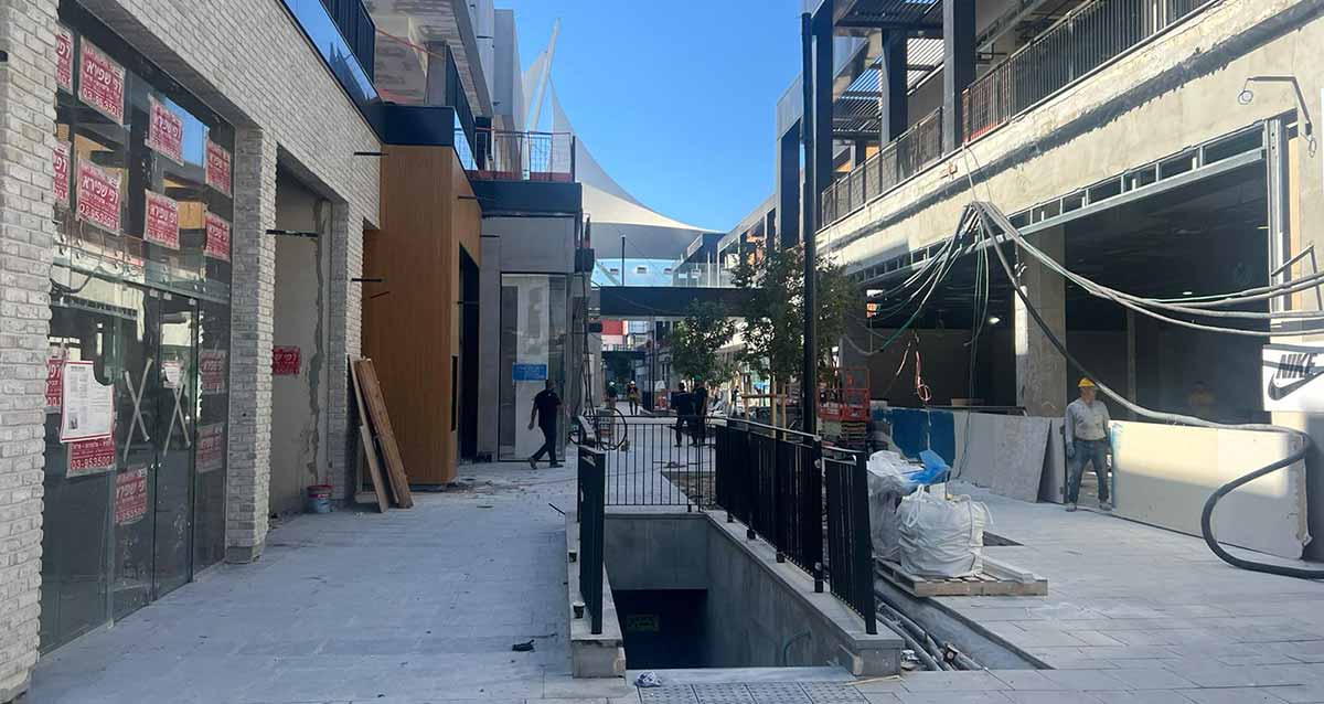 אתר קניון ביג גלילות. תמונה של רחבה בין שני בניינים בשלבי בנייה. באמצע הרחבה מדרגות לחנייה והירידה מגודרת בגדר מתכת שחורה. מספר פועלים ברחבה.