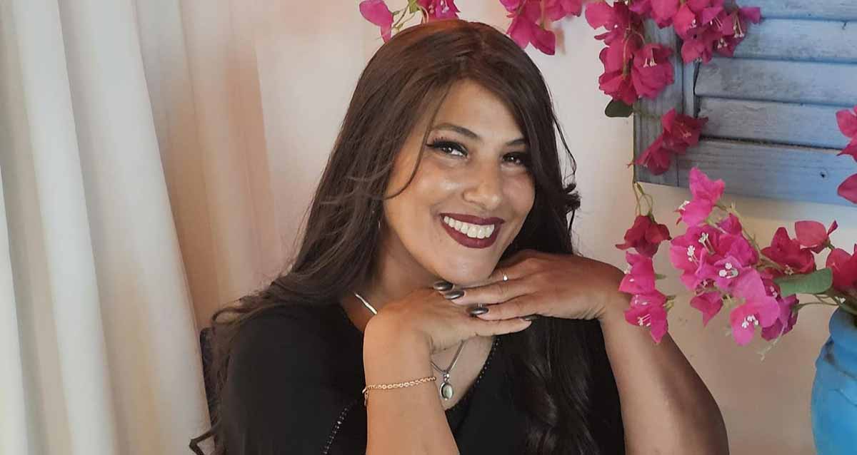 דניאלה חשטא במשרד בצילום פנים, מחייכת, שיער ארוך ושתי ידיה מתחת לסנטר. בצד ימין צמח עם פרחים ורודים אדומים. בצד שמאל וילון בהיר.