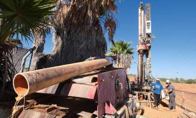 שיקום באר "אל על". צילום של מכונת קידוח עם מנוע בראשו, והוא מסובב מוט מתכת עם גליל שיוצר את הבור. שני עובדים ליד מכונת הקידוח. בצד שמאל מונח צינור ארוך בצורה אופקית על מתקן ממתכת. בצד שמאל עצי דקל.