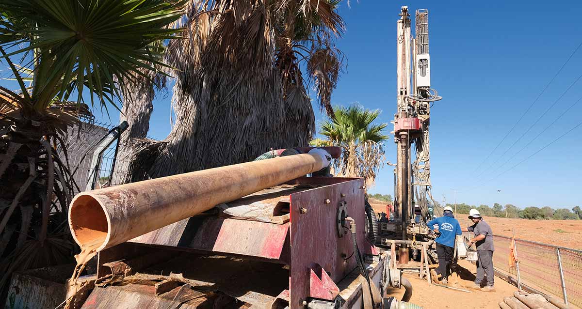 שיקום באר "אל על". צילום של מכונת קידוח עם מנוע בראשו, והוא מסובב מוט מתכת עם גליל שיוצר את הבור. שני עובדים ליד מכונת הקידוח. בצד שמאל מונח צינור ארוך בצורה אופקית על מתקן ממתכת. בצד שמאל עצי דקל.