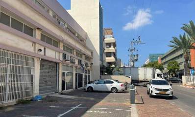 החרושת 12 כפר סבא. בצד שמאל של התמונה בניין של תעשייה ומלאכה בן שלוש קומות. בקומת הקרקע חנויות עם תריסים ושערי מתכת. רובן סגורות. בחזית של הבניין חניות לכלי רכב. חנייה אחת בצד ימין עם מחסום אופקי. מימינו חונה מכונית אחת. מכונית נוספת חונה על המדרכה לפני המחסום. אחרי הבניין בן שלוש הקומות מחובר אליו בניין גבוה עם חלונות לחזית ואחריו עוד בניין. בצד ימין של התמונה בניין תעשייה ומלאכה בן ארבע קומות עם חזית אפורה וקירות חומים בשני הצדדים. הכביש חסום עם משאית היוצאת מאחורי הבניין בצד שמאל. עמוד חשמל של מתח גבוה במדרכה בצד ימין ברקע.