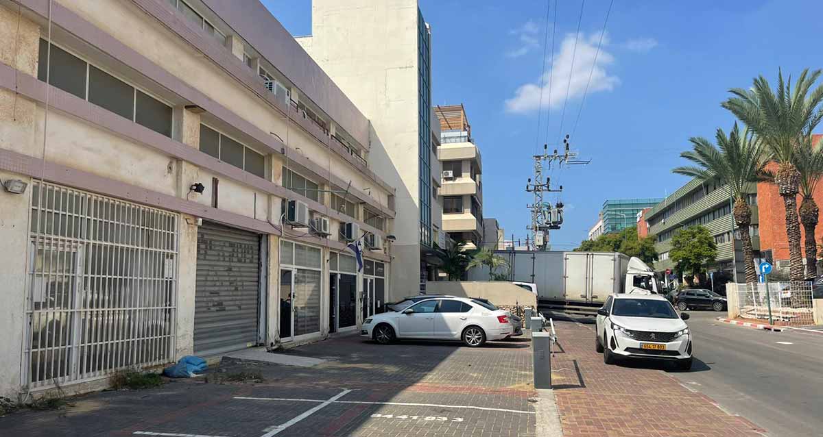 החרושת 12 כפר סבא. בצד שמאל של התמונה בניין של תעשייה ומלאכה בן שלוש קומות. בקומת הקרקע חנויות עם תריסים ושערי מתכת. רובן סגורות. בחזית של הבניין חניות לכלי רכב. חנייה אחת בצד ימין עם מחסום אופקי. מימינו חונה מכונית אחת. מכונית נוספת חונה על המדרכה לפני המחסום. אחרי הבניין בן שלוש הקומות מחובר אליו בניין גבוה עם חלונות לחזית ואחריו עוד בניין. בצד ימין של התמונה בניין תעשייה ומלאכה בן ארבע קומות עם חזית אפורה וקירות חומים בשני הצדדים. הכביש חסום עם משאית היוצאת מאחורי הבניין בצד שמאל. עמוד חשמל של מתח גבוה במדרכה בצד ימין ברקע.