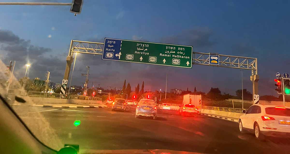 צומת הכפר הירוק רמת השרון, שלט הכוונה על גשר ממתכת, בצד ימין השלט בצבע ירוק הכוונה לרמת השרון והרצליה, ובצד שמאל הכוונה לכביש 5 לחיפה ברקע כחול. מתחת לשלט מכוניות עם אורות דלוקים.