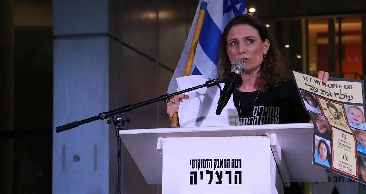 עידית אלכסנדרוביץ נואמת בהפגנה בהרצליה. מחזיקה בידה עלון ובו כתוב שלח את עמי LET MY PEOPLE GO ובו תמונות של 6 של חטופים ושל יולי אדלשטיין כאסיר ציון