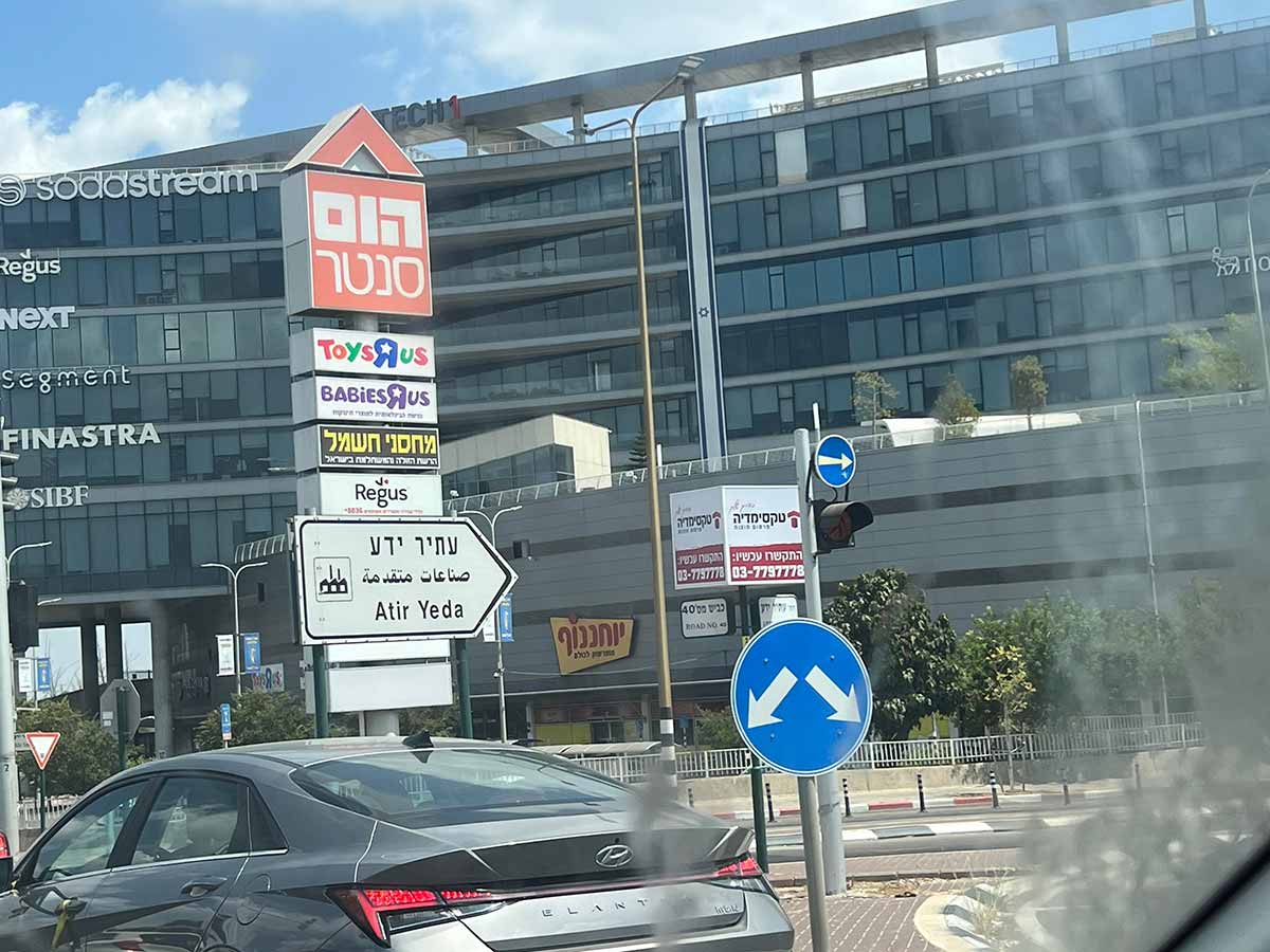 אזור התעשייה עתיר ידע בכפר סבא, בניין בן 7 קומות, בו הקירות החיצוניים מצופים בלוחות זכוכית. לפניו שלט גדול ועליו שמות עסקים: הום סנטר, טויס אר אס ובייבי אר אס באנגלית, מחסני חשמל, רגוס באנגלית. מימין שלט של יוחננוף ומשרד של תחנת מוניות טקסימדיה.