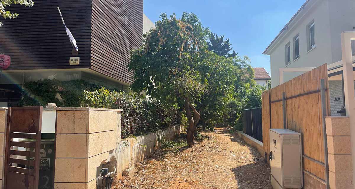 כצנלסון 42 כפר סבא. שני בתים פרטיים וביניהם שביל לא מרוצף התחום בגדרות בטון של שני המגרשים. בצד ימין על השביל ארון של הוט וממול שעון המים של הבית השמאלי. בצד ימין גדר של קני סוף של הבית הימני. הבית משמאל מצופה עם לוחות חומים בקירות הקדמי והצדדי, והחלק האחורי של הבית מרוצף באריחים בהירים. דגל ישראל תלוי על הקיר הכהה. בצד שמאל של השביל בין הבתים עצים גדולים.