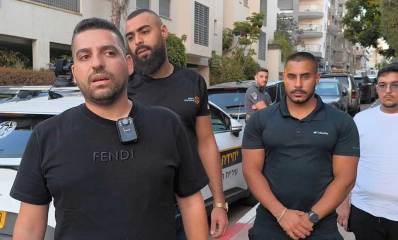 מספר גברים, חלקם שומרים גברתניים עומדים ברחוב לצד רכב של יחידת אכיפה עירונית. אחד מהם לובש חולצה שחורה עם מצלמת גוף מחוברת לחולצתו. שני גברים נוספים לובשים חולצות שחורות ועומדים ברקע, תלמיד ישיבה נוסף עם חולצה לבנה וכיפה עומד בצד ימין. הרקע כולל מכוניות חונות ובנייני מגורים