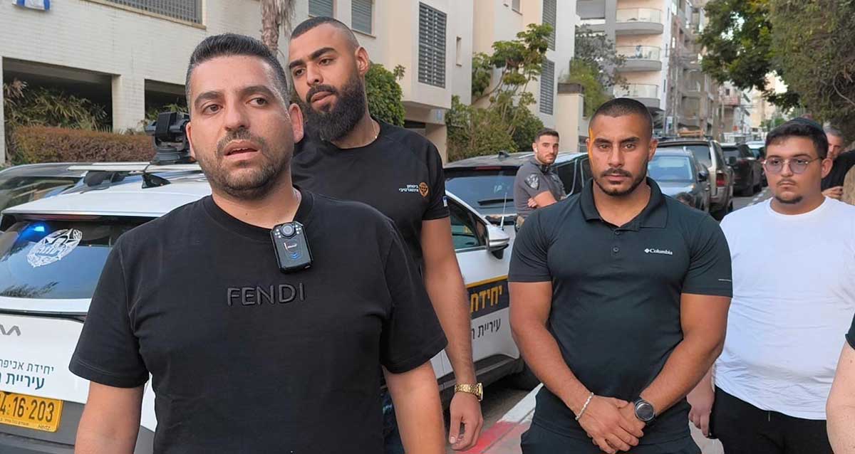 מספר גברים, חלקם שומרים גברתניים עומדים ברחוב לצד רכב של יחידת אכיפה עירונית. אחד מהם לובש חולצה שחורה עם מצלמת גוף מחוברת לחולצתו. שני גברים נוספים לובשים חולצות שחורות ועומדים ברקע, תלמיד ישיבה נוסף עם חולצה לבנה וכיפה עומד בצד ימין. הרקע כולל מכוניות חונות ובנייני מגורים