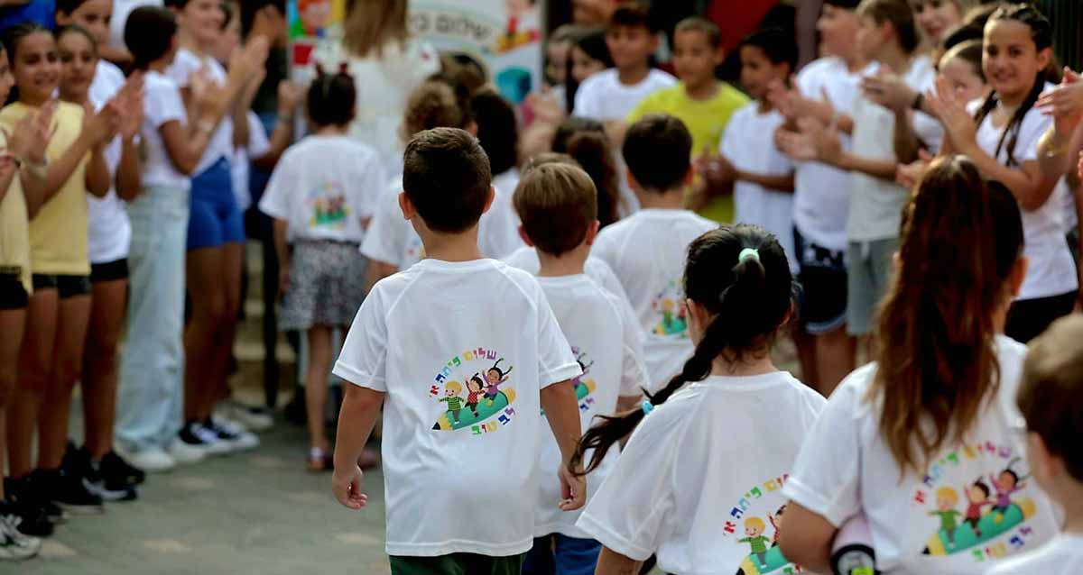 פתיחת שנת הלימודים בבית הספר לב טוב. תלמידים של כיתות גבוהות מקבלים את פניהם של תלמידי כיתה א', הלבושים בחולצות לבנות של בית הספר ובגב החולצה ציור של שלושה ילדים רוכבים על קלמר גדול וכיתוב: שלום כיתה א', לב טוב