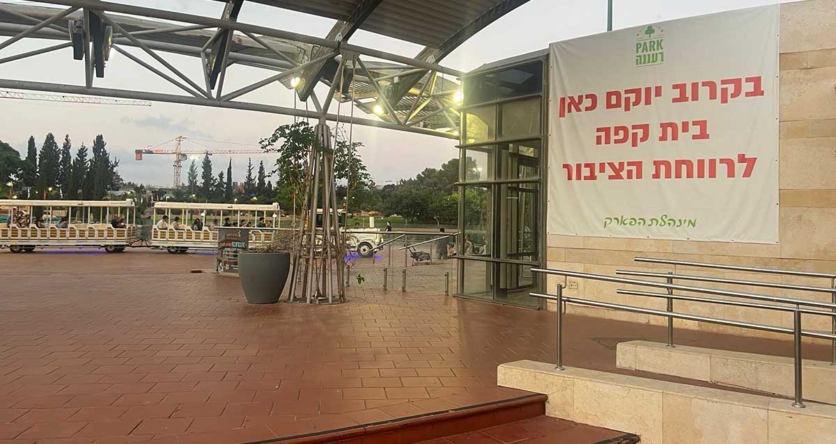 צילום של פארק רעננה עם רכבת הפארק מסיעה אנשים ומנוף ברקע. שלט ענק על הקיר של בית הקפה עם הכיתוב: prark רעננה. בקרוב יוקם כאן בית קפה לרווחת הציבור. מינהלת הפארק