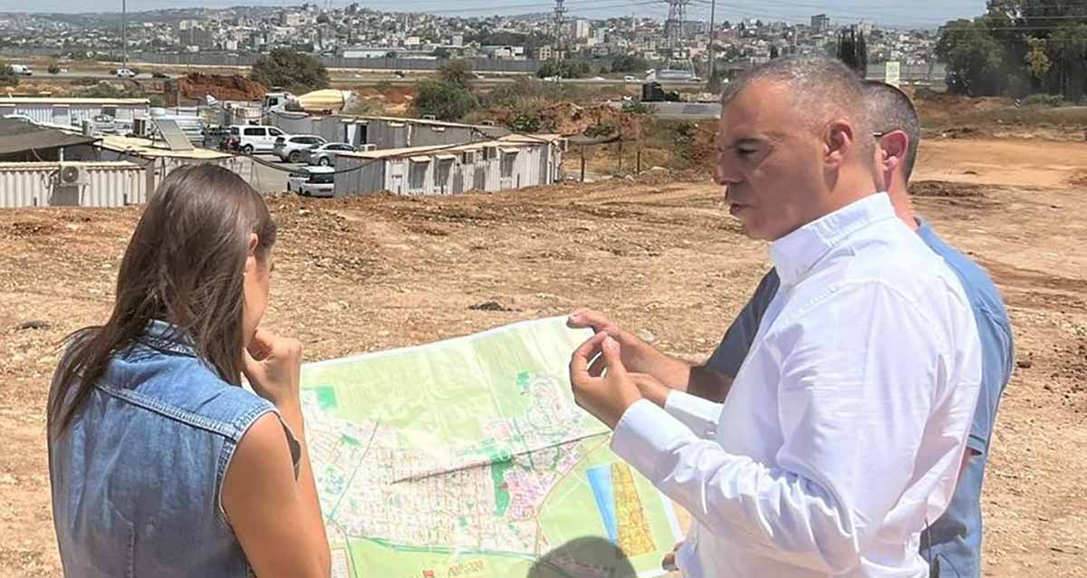 ראש העיר רפי סער עם תוכנית קריית הצעירים בכפר סבא. ראש העיר רפי סער עומד בשטח פתוח עם עוד שני אנשים האוחזים במפה של הקרייה המתוכננת בשטח זה. בקצה השטח מוסך לכלי רכב. וברקע בנייני העיר.