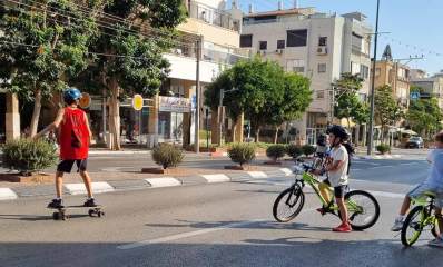 ילדים על אופניים וסקטבורד על הכביש. שני ילדים עם קסדות, וילד אחד עם אופניים ללא קסדה. אין מכוניות בכביש, וברקע בנייני מגורים בני 3 עד 5 קומות לאורך הרחוב.