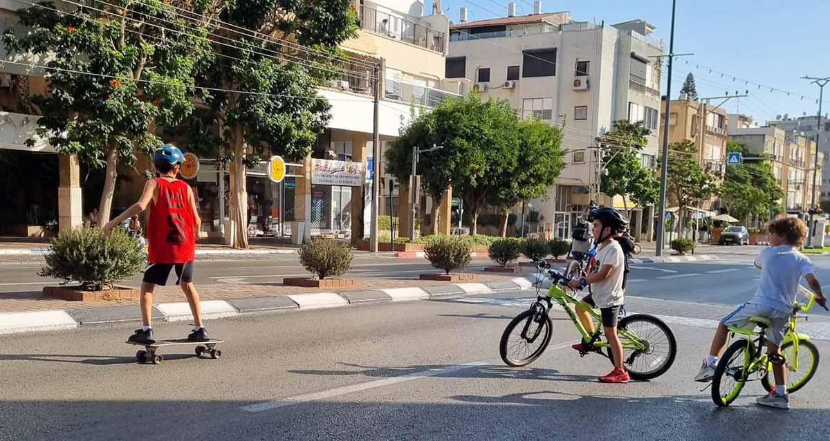 ילדים על אופניים וסקטבורד על הכביש. שני ילדים עם קסדות, וילד אחד עם אופניים ללא קסדה. אין מכוניות בכביש, וברקע בנייני מגורים בני 3 עד 5 קומות לאורך הרחוב.