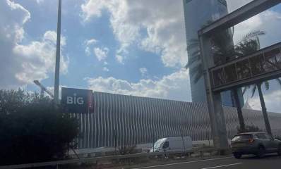 מתחם ביג בגלילות בבנייה. תמונה מהכביש אל החומה המקיפה את המתחם. קיר בטון עם פסים לכל אורך החומה. מימין מגדל משרדים. ולידו גשר לשילוט מעל הכביש.