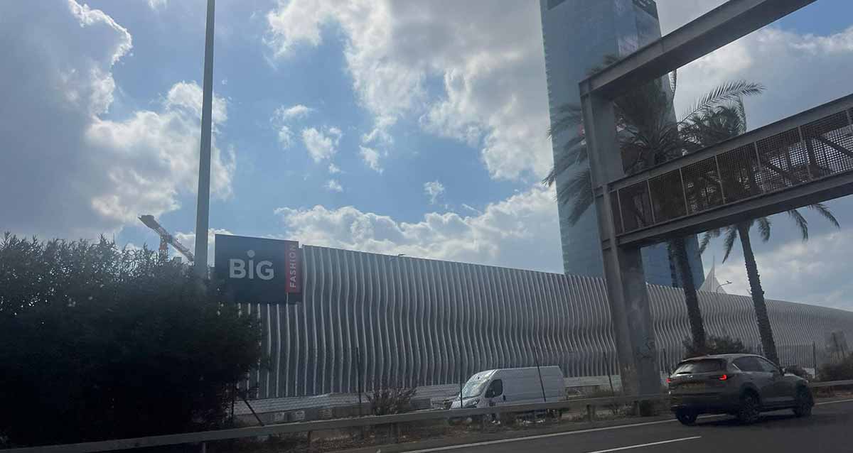 מתחם ביג בגלילות בבנייה. תמונה מהכביש אל החומה המקיפה את המתחם. קיר בטון עם פסים לכל אורך החומה. מימין מגדל משרדים. ולידו גשר לשילוט מעל הכביש.