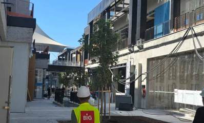 מתחם ביג בגלילות בבנייה. מימין ומשמאל בניינים של שתי קומות וביניהן רחבה. ברחבה כניסה לירידה לחניון. בתחתית התמונה גב של פועל עם חולצה צהובה וכיתוב באנגלית: BIG.