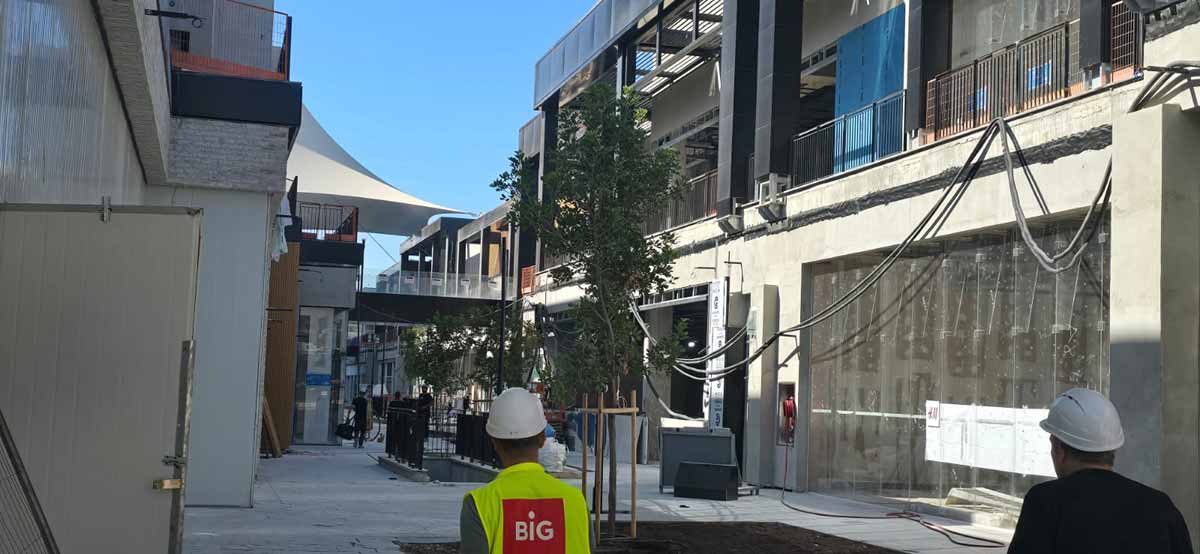 מתחם ביג בגלילות בבנייה. מימין ומשמאל בניינים של שתי קומות וביניהן רחבה. ברחבה כניסה לירידה לחניון. בתחתית התמונה גב של פועל עם חולצה צהובה וכיתוב באנגלית: BIG.