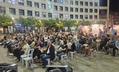 מציינים בהרצליה שנה למלחמה. קהל יושב על כסאות פלסטיק לבנים בטקס ברחבת העירייה.