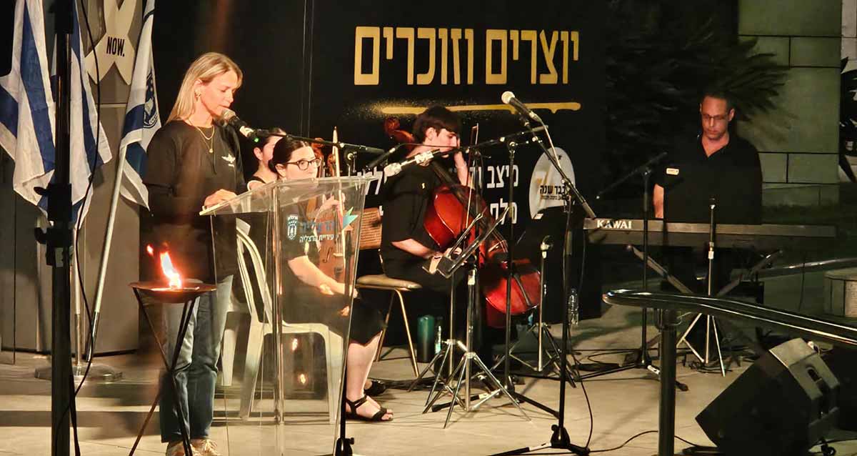 מציינים בהרצליה שנה למלחמה. בחורה נואמת על הבמה לפניה מיקרופון ודוכן נואמים שקוף. מימינה נגן קלידים, נגן גיטרה ונגנית צ'לו. ברקע הכיתוב: יוצרים וזוכרים.
