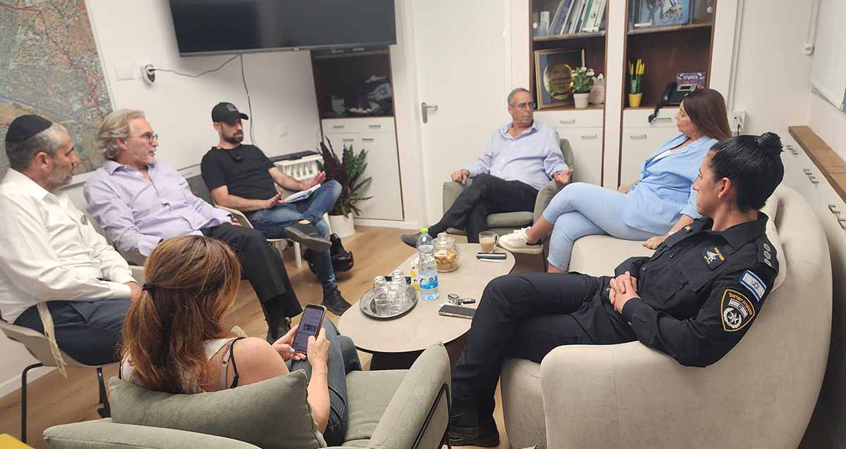 פגישת ראש העיר עם מתנדבים. מסביב לשולחן אליפטי יושבים ראש העיר ומתנדבים על כורסאות וכסאות. על השולחן בקבוק מים, כוסות וצנצנת עוגיות.