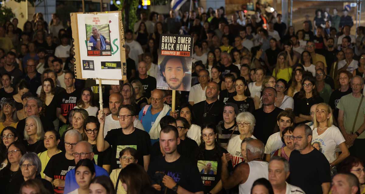 טקס זיכרון 7.10 בהרצליה. המוני נשים וגברים עומדים בצפיפות. שניים מהמשתתפים בטקס אוחזים בידיהם מוטות עם תמונות של חטופים. שלט אחד עם כיתוב באנגלית: BRING OMER BACK HOME ותמונה גדולה של עומר. השני אוחז שלט גדול עם תמונה של חטוף.