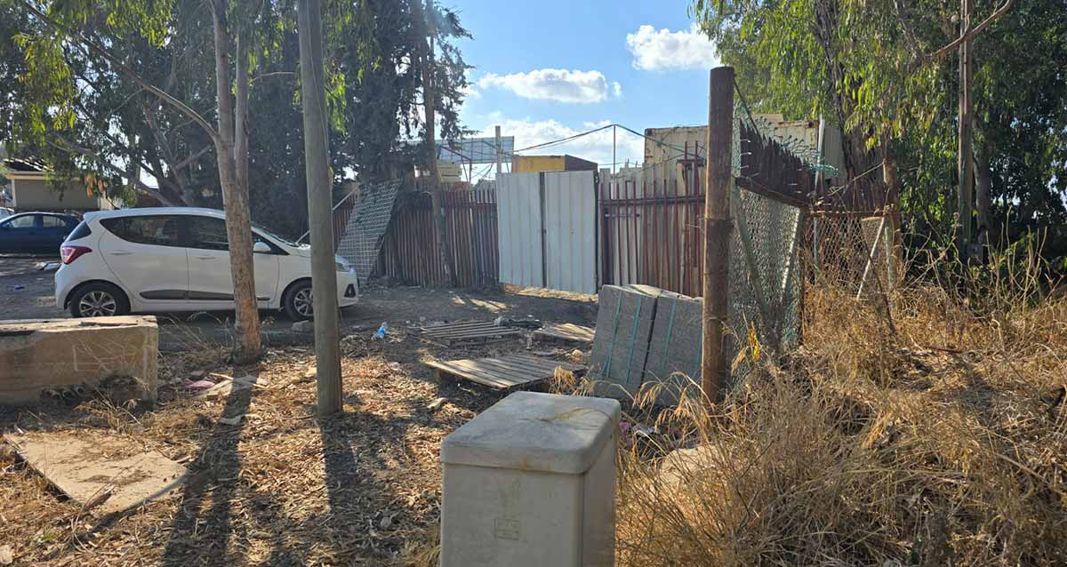 חורשת הנ"מ בהרצליה. ברקע גדר של מתחם סגור ושער לבן. מכונית לבנה ליד השער. בצד ימין זרוקים ארונות. בצד שמאל שתי מכוניות נוספות.