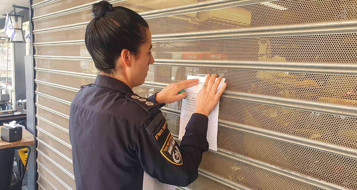 שוטרת תולה צו סגירה, דף נייר, על תריס סגור של הכניסה למסעדה.