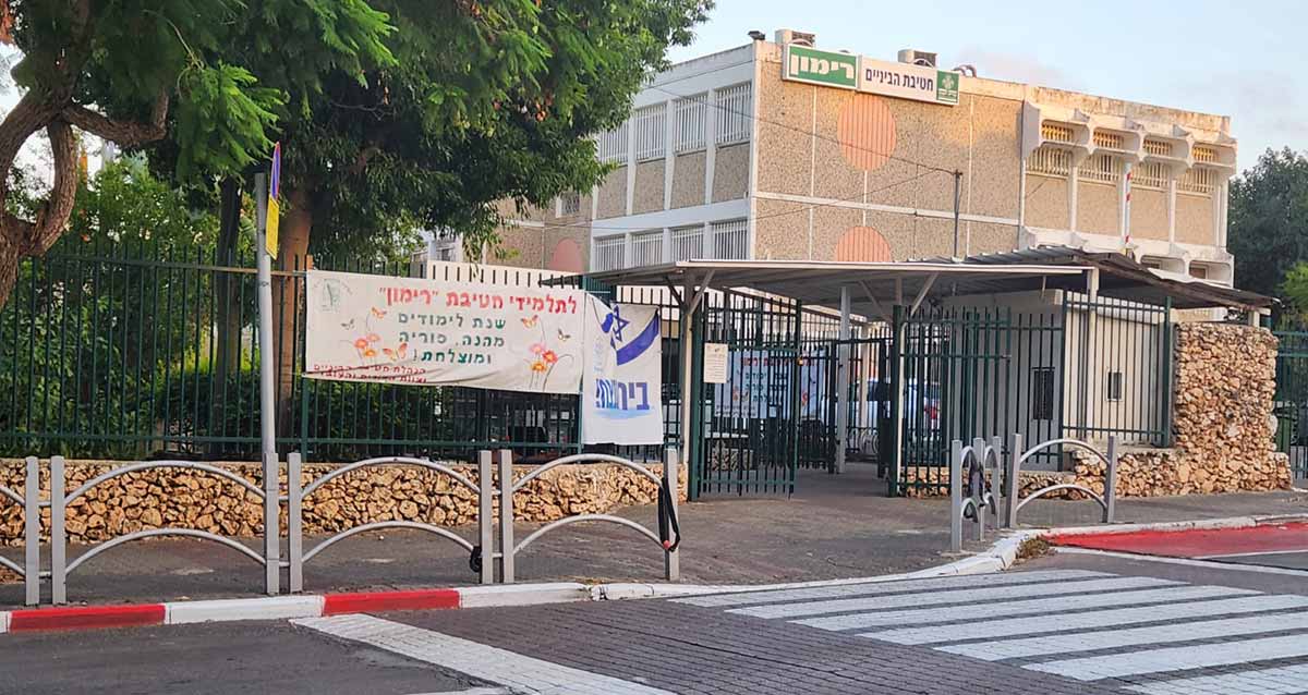 חטיבת הביניים רימון ברעננה. תמונה של בניין בית הספר שצולמה מהכביש שלפני בית הספר. מעבר חצייה להולכי רגל אל שערי בית הספר. חומה מבטון ואבנים מסביב לבית הספר וגדר מתכת על החומה. שערי מתכת של בית הספר. הבניין בן שלוש קומות, על הקומה העליונה שלט גדול, מימין סמל, באמצע על רקע לבן הכיתוב: חטיבת הביניים, ומשמאל על רקע ירוק באותיות גדולות הכיתוב: רימון. על גדר בחזית תלוי שלט מבד לבן ועליו הכיתוב: לתלמידי חטיבת "רימון" שנת לימודים מהנה, פוריה ומוצלחת.
