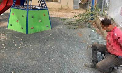 שוהה בלתי חוקי נעצר בהרצליה. בגן משחקים יושב על כסא שב"ח אזוק ופניו מטושטשות. הוא לבוש מכנסיים חומים וחולצה אדומה. במרכז התמונה מגלשה ומתקן עליה לראש המגלשה.