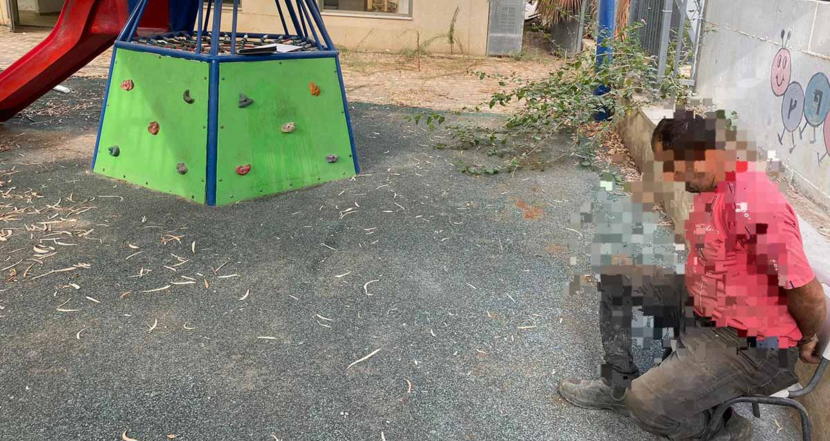 שוהה בלתי חוקי נעצר בהרצליה. בגן משחקים יושב על כסא שב"ח אזוק ופניו מטושטשות. הוא לבוש מכנסיים חומים וחולצה אדומה. במרכז התמונה מגלשה ומתקן עליה לראש המגלשה.