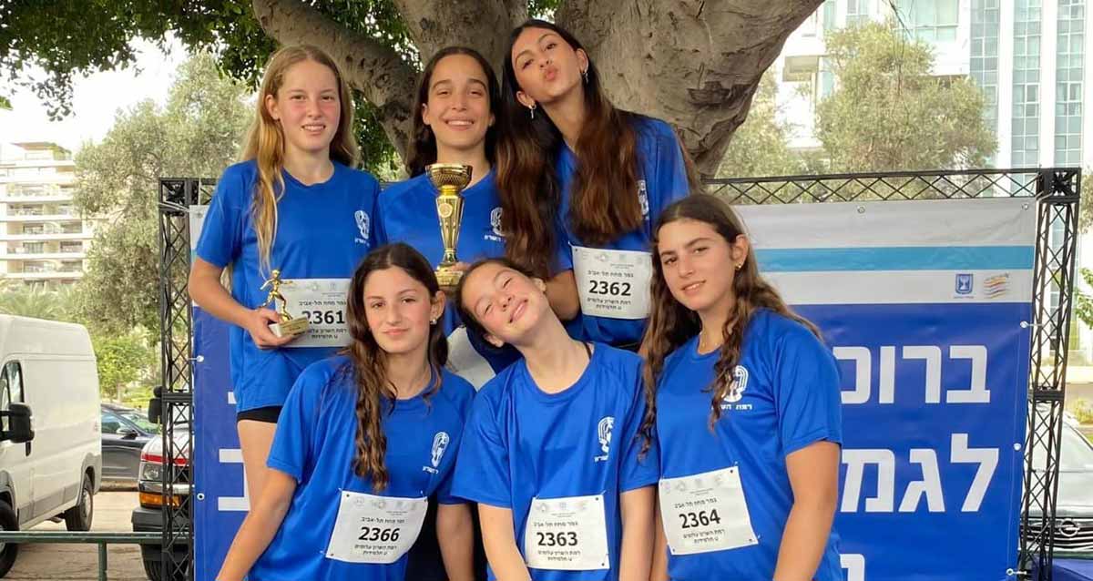 נבחרת הריצה של תלמידות עלומים. שש בנות בתלבושת ספורט ובחולצה סמל של רמת השרון. על החולצה מחובר שלט לבן עם המספר של כל אחת. אחת הבנות מחזיקה גביע.