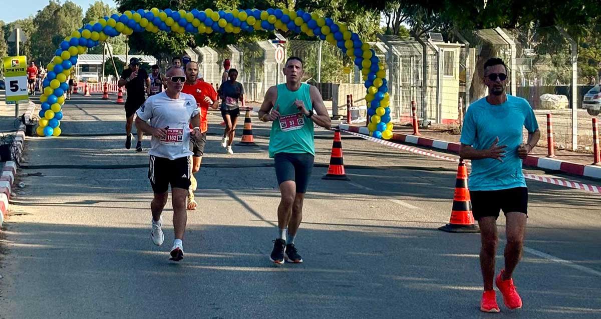 רצים במרוץ אייל 2024 ברמת השרון, המסלול על כביש מסומל בקונוסים זוהרים ובסר סימון, מאחורי הרצים שלט 5 ק"מ ושער בלונים צהובים כחולים ענק