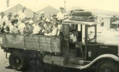 קיבוץ רמת הכובש בבתחילת הדרך. על משאית יושבים ועומדים בחורות ובחורים. מאחוריהם נראים מבנים של הקיבוץ, עם גגות משופעים.