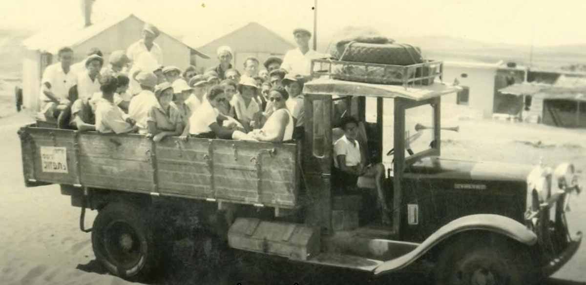 קיבוץ רמת הכובש בבתחילת הדרך. על משאית יושבים ועומדים בחורות ובחורים. מאחוריהם נראים מבנים של הקיבוץ, עם גגות משופעים.