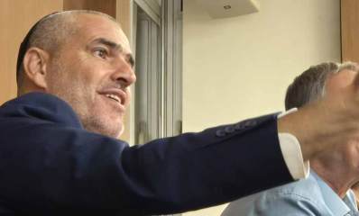 יעקב קורצקי מניף ידו הימנית קדימה. צילום פלג גוף עליון. לבוש ז'קט כחול וחולצה לבנה.