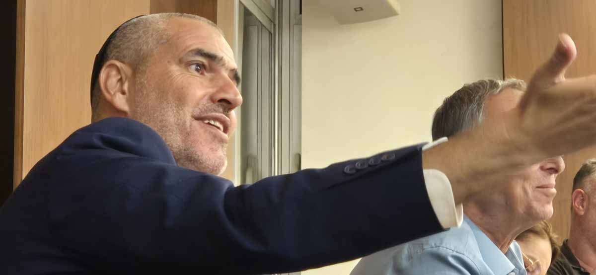 יעקב קורצקי מניף ידו הימנית קדימה. צילום פלג גוף עליון. לבוש ז'קט כחול וחולצה לבנה.