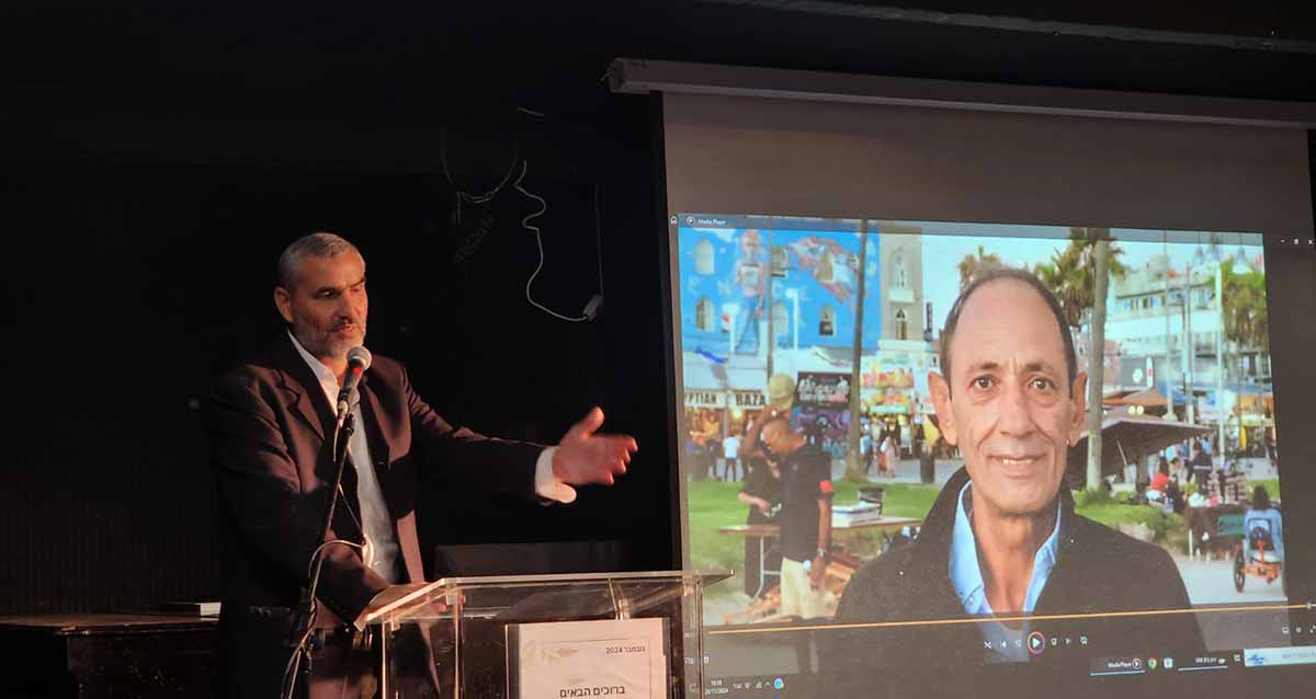 סגן ראש העיר יעקב קורצקי עומד מאחורי דוכן נואמים עם מיקרופון, ומברך את איציק כהן מנהל מחלקת התברואה שפרש, ותמונתו מוקרנת על מסך.