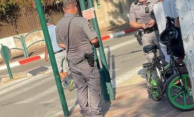 אכיפת קנסות קורקינטים ברמת השרון. שני שוטרים ליד קורקינט על מדרכה. בכביש מעבר חצייה והולך רגל חוצה את הכביש. משמאל גדר עץ של בית.